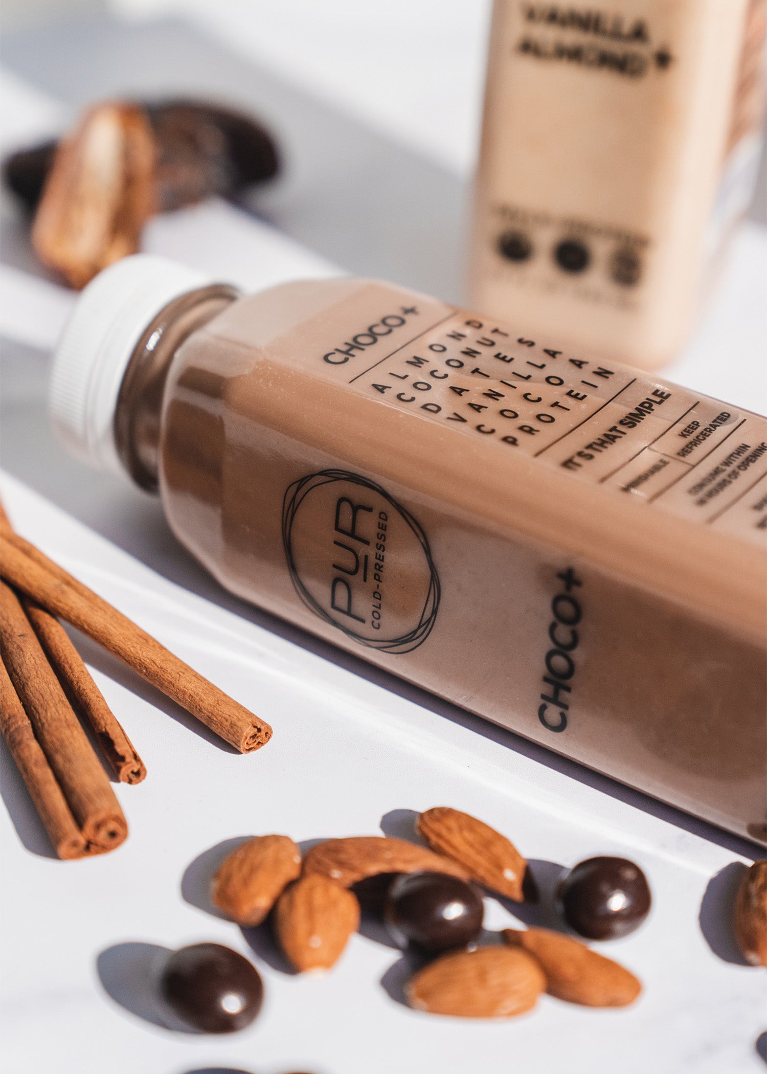Choco+ protein bottle with vanilla almond flavor on a white surface with cinnamon sticks and almonds.