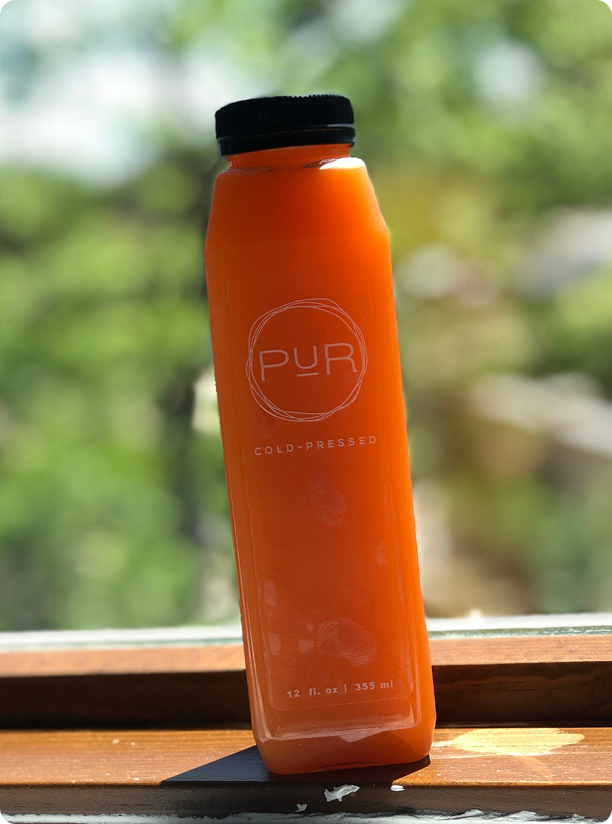 Single PUR Cold Pressed sunny citrus juice bottle backlit on a windowsill.