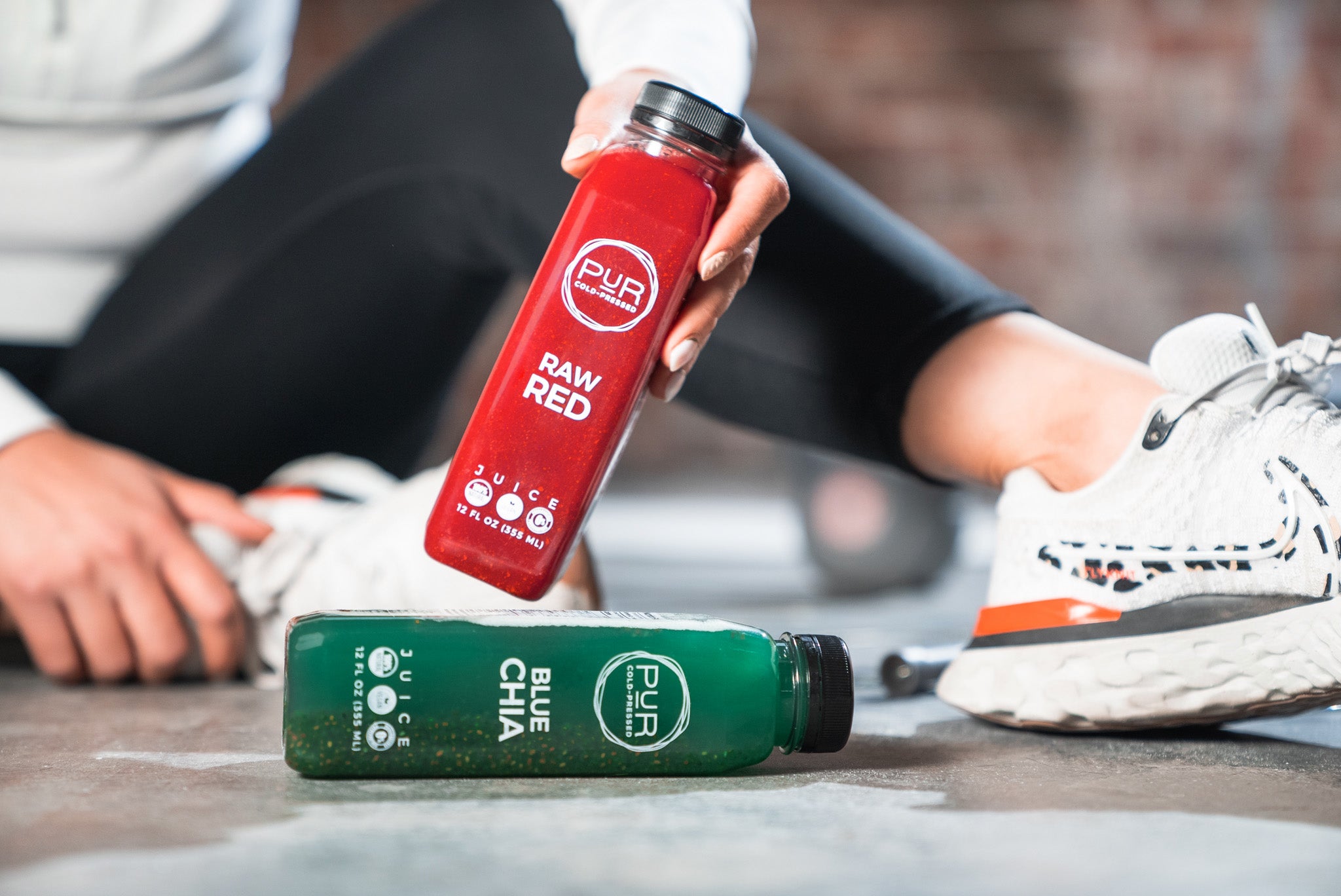 Hand picking up a PUR Raw Red bottle above a PUR Blue Chia bottle on a gym floor, with white running shoes in the background.