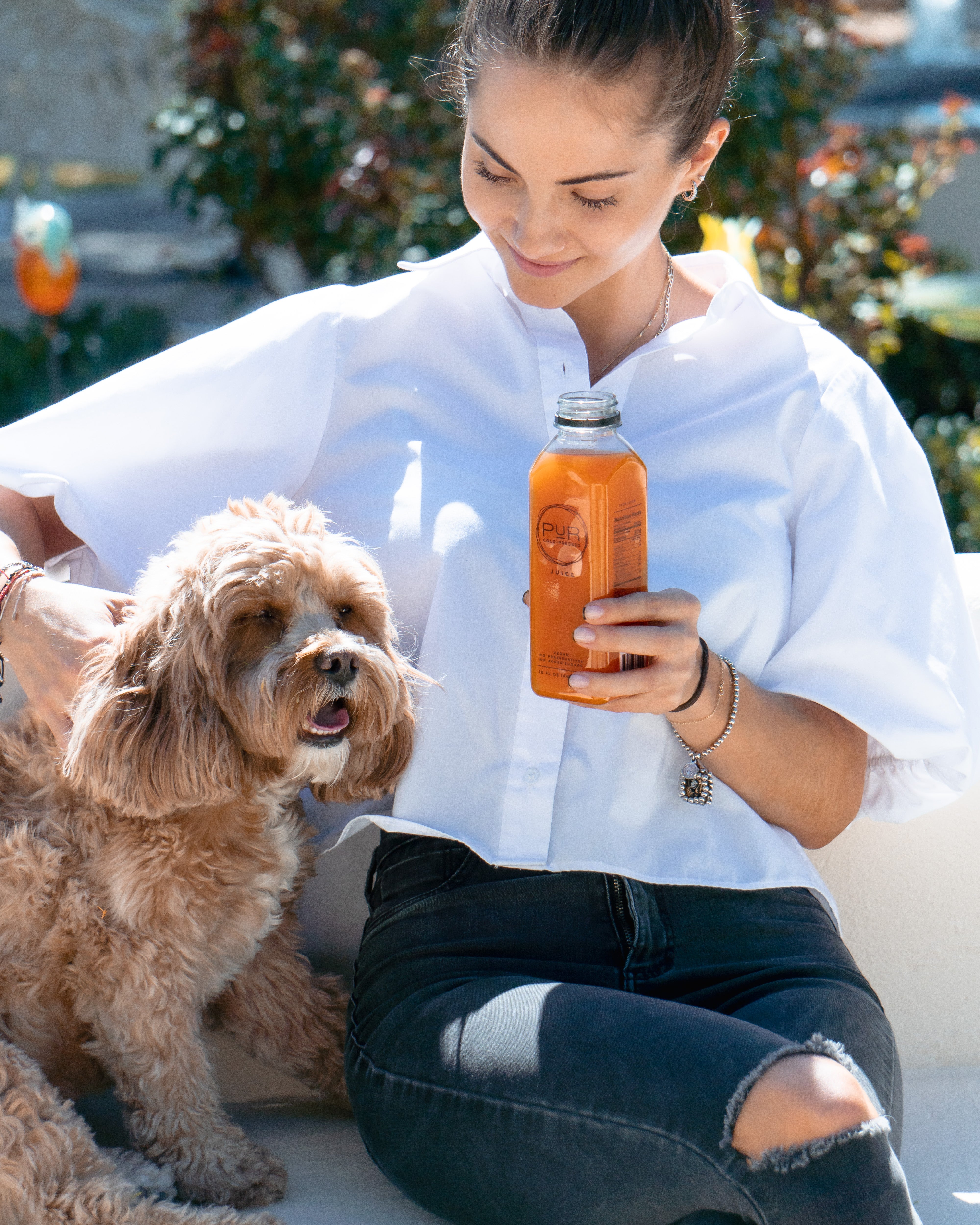 Woman sitting outdoors with her dog, holding a bottle of PUR Cold-Pressed juice.