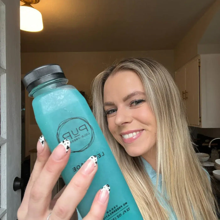 Person smiling while holding a bottle of blue PUR Cold-Pressed Blue Majik lemonade.