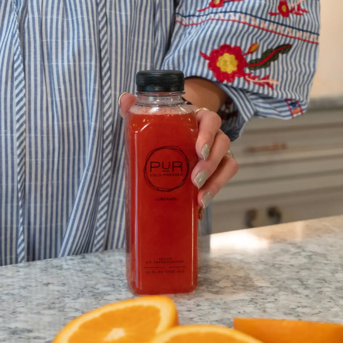 Person holding a bottle of PUR Cold-Pressed red lemonade on a kitchen counter.
