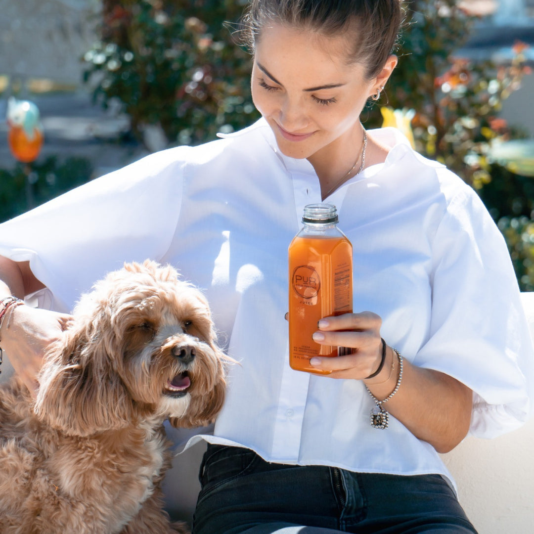 Woman smiling outdoors holding PUR Cold Pressed sunny citrus juice while petting her dog.