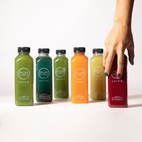 Hand reaching for PUR Cold Pressed juices in assorted flavors including Greens, Sunny Citrus, and Raw Red.