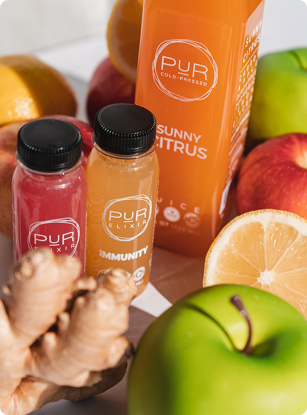 PUR Sunny Citrus with Immunity and red elixir shots among apples, orange slices and ginger.